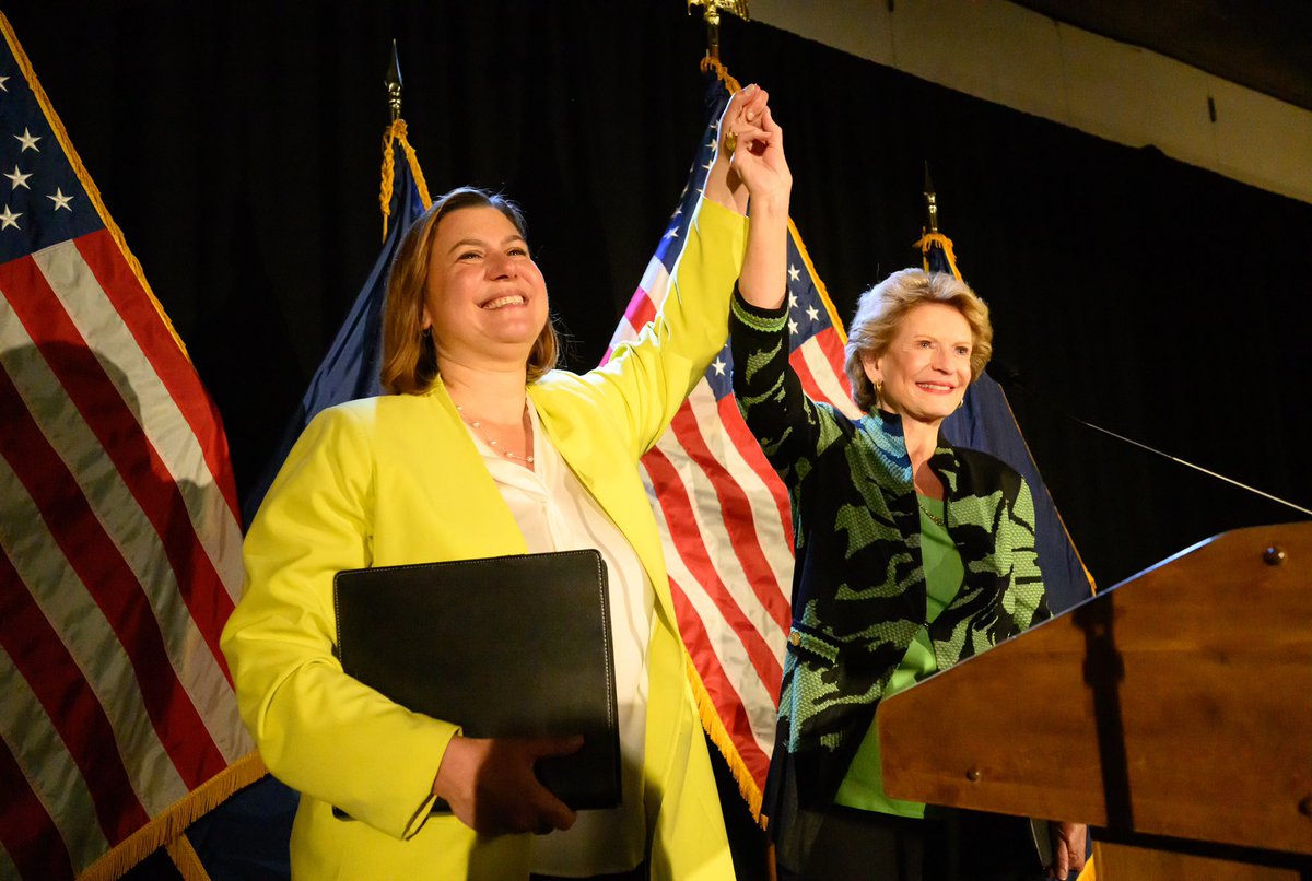 Debbie Stabenow with Senator-elect Elissa Slotkin holding raised arms in front of flags at a podium.