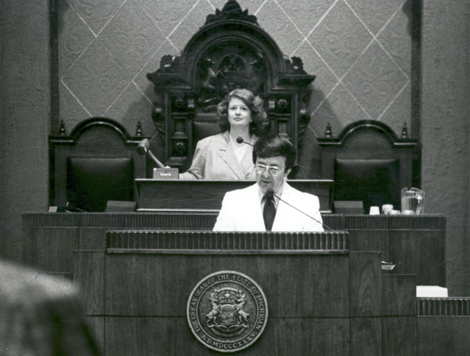 Debbie Stabenow presides over Michigan State House with gavel, sitting above a male speaker.