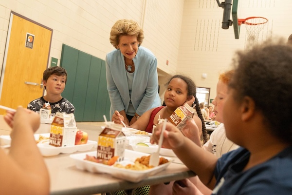 Senator Stabenow talking to four children who are sitting at a table eating a school lunch