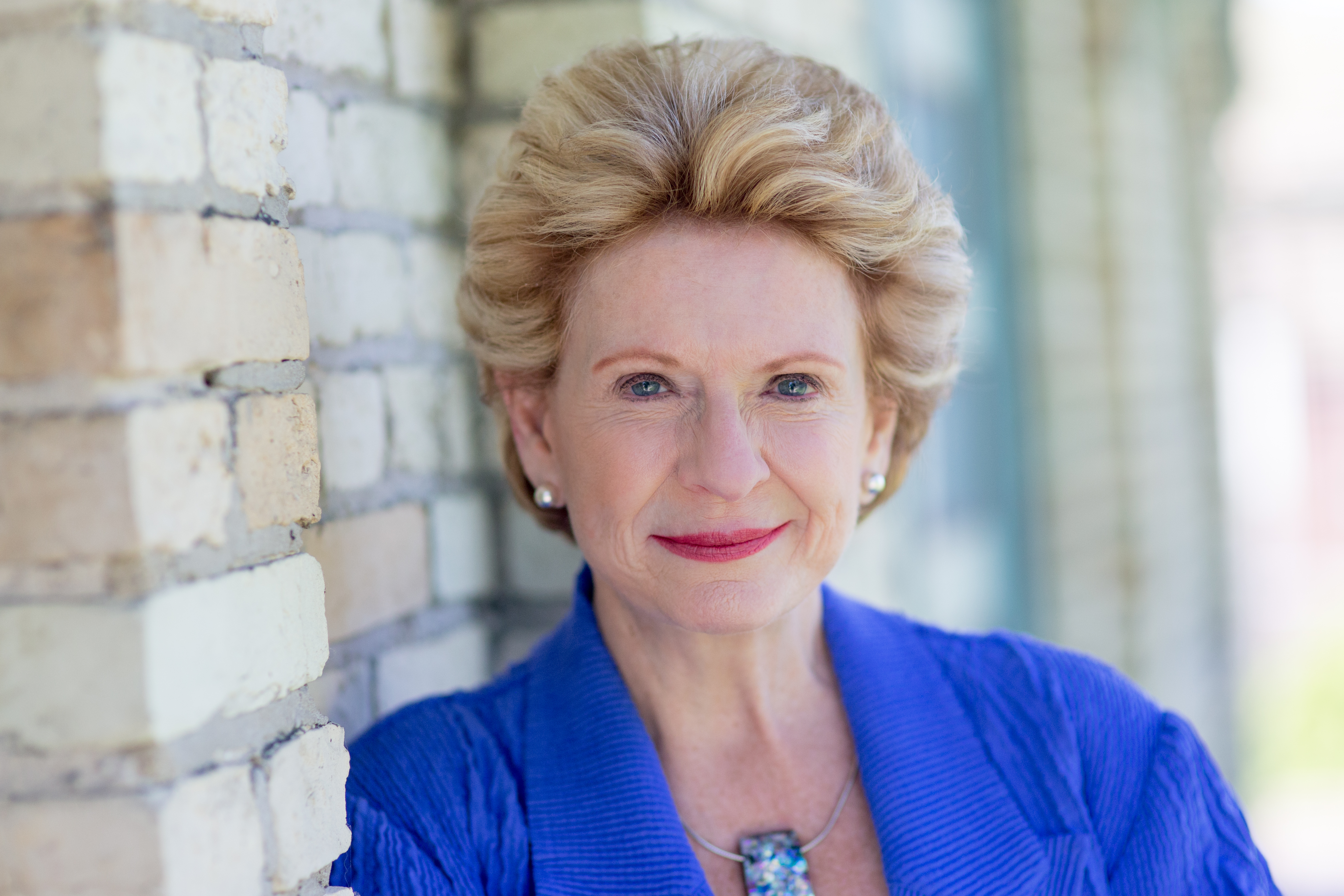 High-resolution photo of Senator Debbie Stabenow in a blue outfit, leaning her shoulder against a brick wall