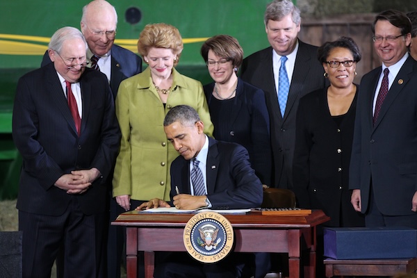 A group of elected officials standing behind President Obama who is seated at a desk signing a piece of paper