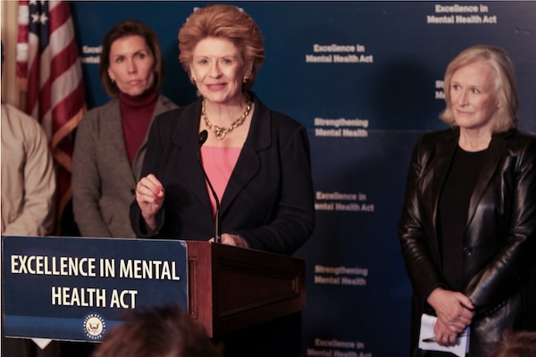 Debbie Stabenow speaks from a podium that has a sign that says Excellence in Mental Health Act while actress Glenn Close stands next to her.