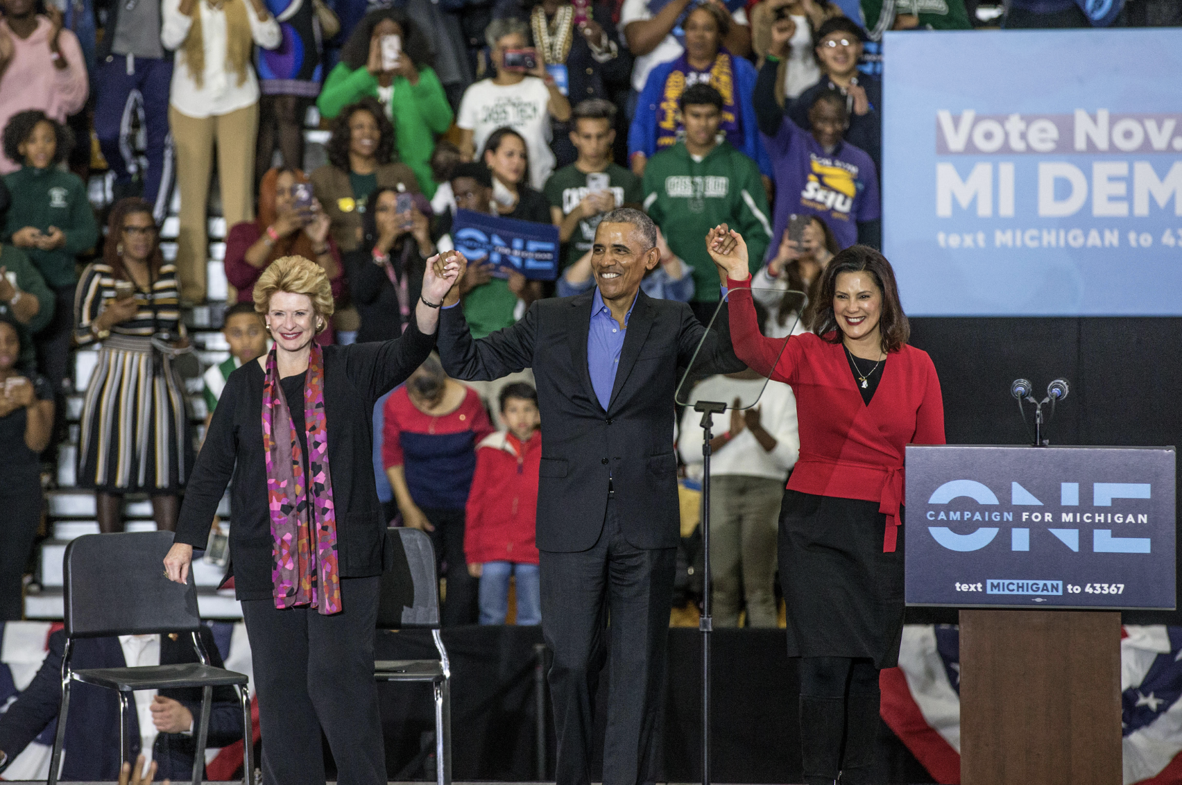 Debbie Stabenow, President Obama and governor Whitmer, with raised hands on a stage in front of a political rally crowd.